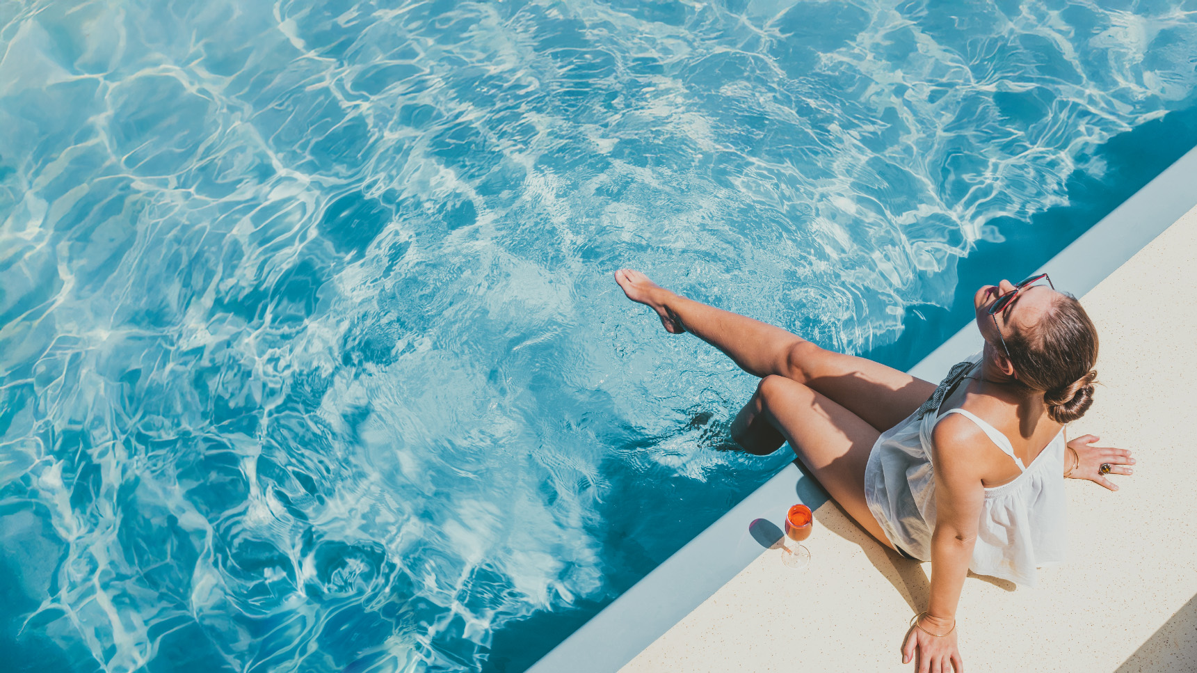 woman sitting by the pool with her feet in the water and a cocktail by her side looking up into the sun