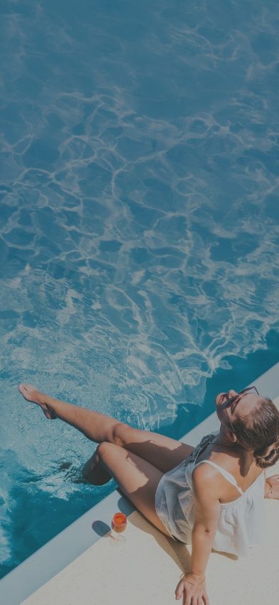 woman sitting by the pool with her feet in the water and a cocktail by her side looking up into the sun