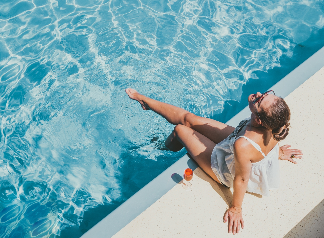 woman sitting by the pool with her feet in the water and a cocktail by her side looking up into the sun