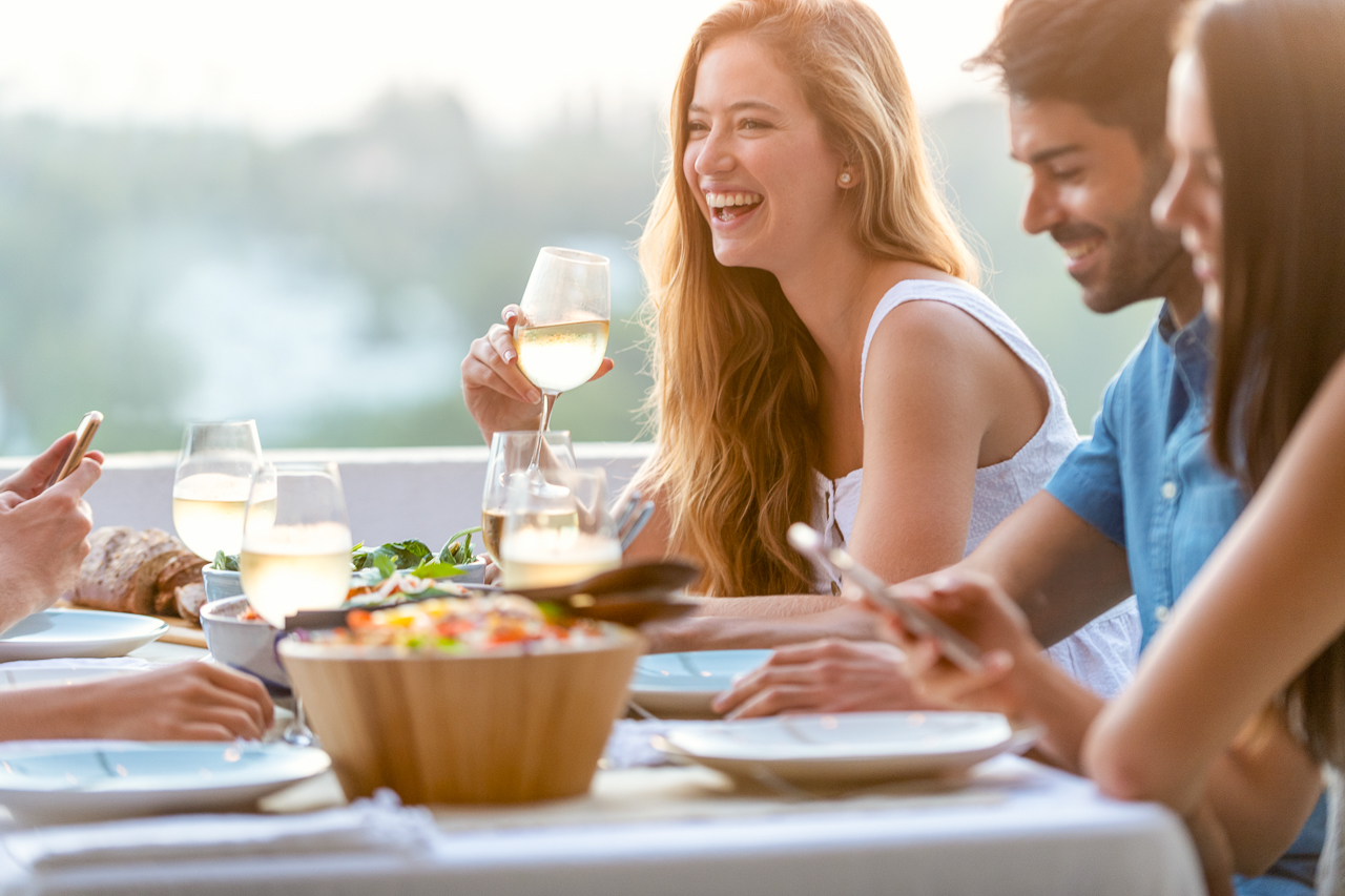 Friends chatting and drinking at a dinner party with wine glasses on the table. There are also bowls of other food and salad on the table. Time is sunset outdoors. There are 2 people using their mobile phones as they celebrate Riversong life.