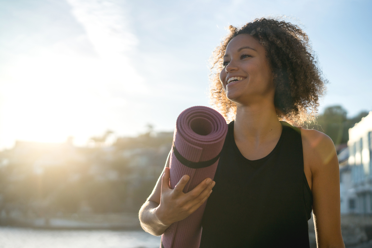Portrait of a fit woman holding a yoga mat at the beach and looking very happy - wellness concepts - Riversong life