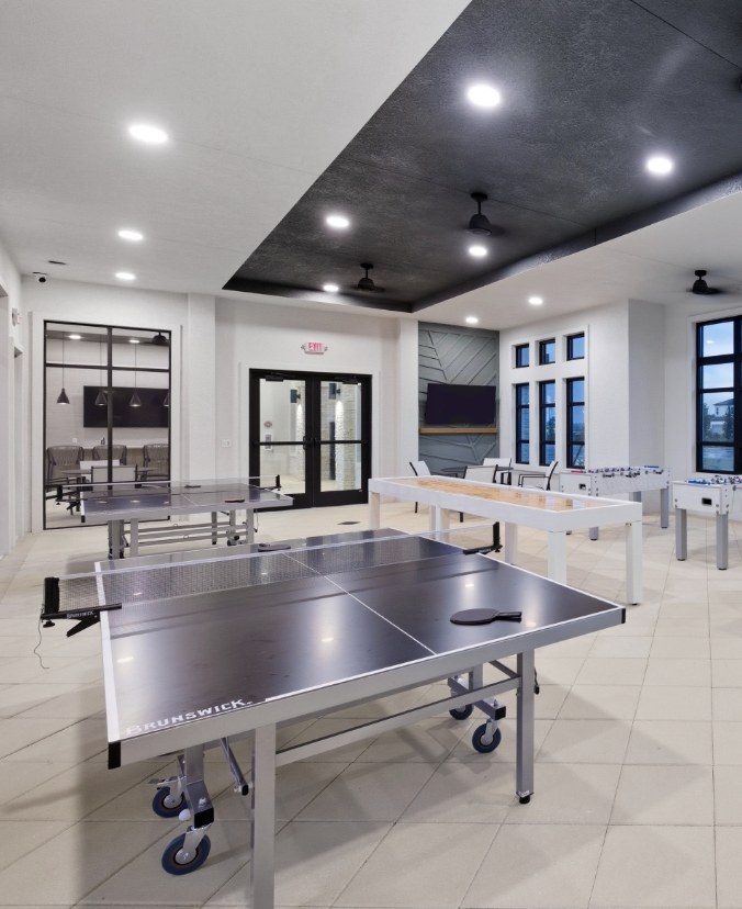 Bright, modern game room at Riversong with multiple ping-pong tables in the foreground, a shuffleboard table and foosball table in the background, and large windows creating an open, recreational space.